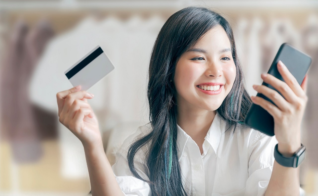 Portrait of happy young asian woman showing credit card and using smartphone while shopping at mall.
