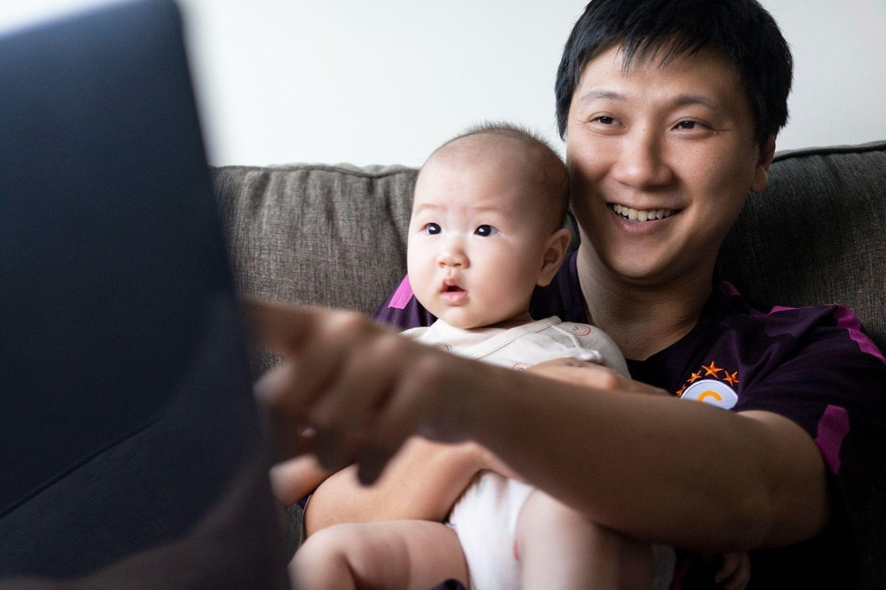 Lim WeiXiang carrying his infant child and pointing to the screen on his laptop.