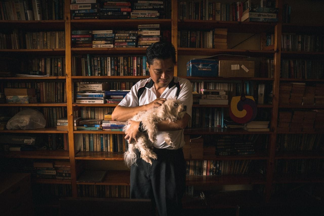 Portait image of Ronald Hee with his dog in his arms, standing in front of a shelf filled with books. 