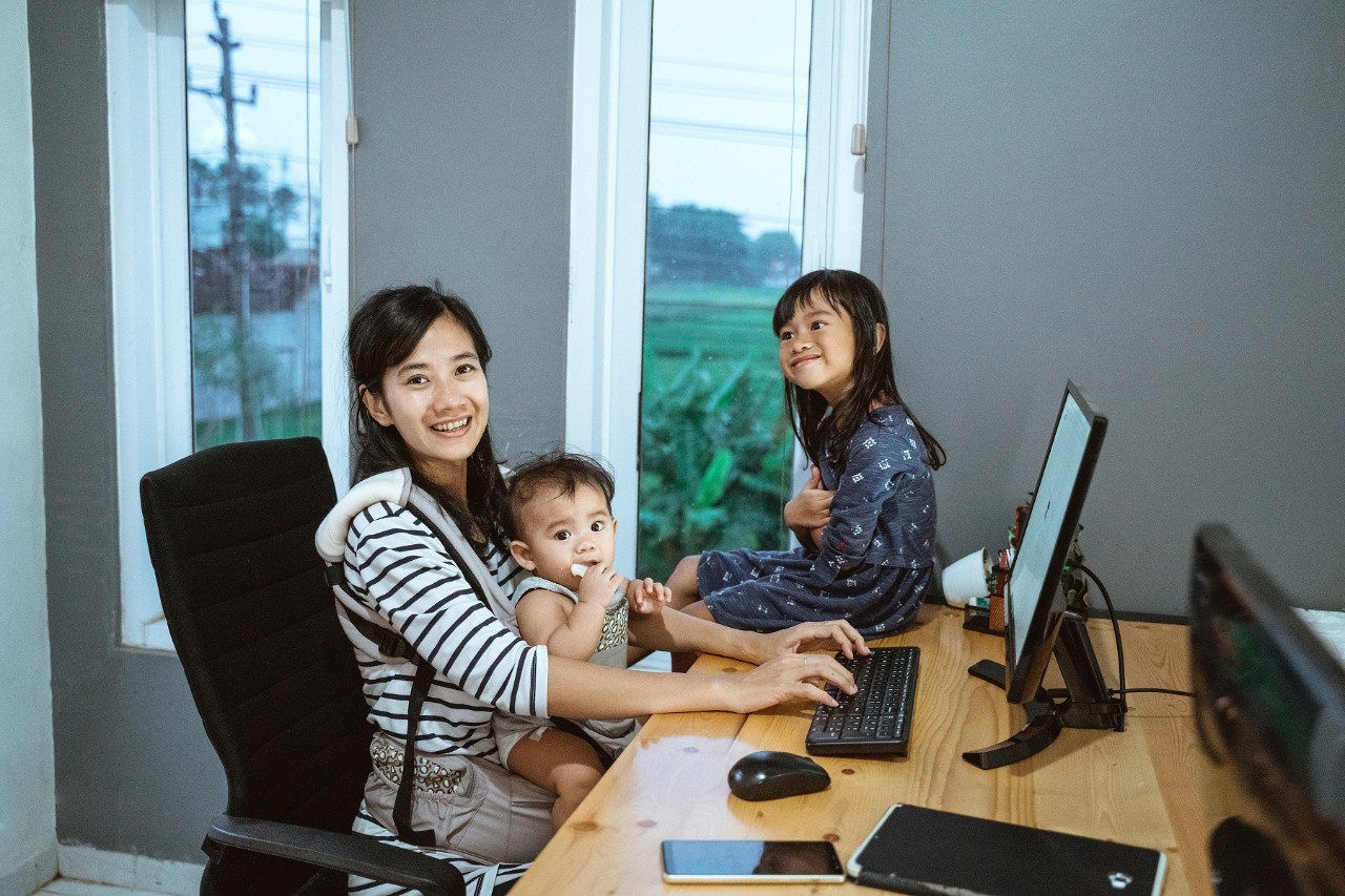 Mother with two children and one strapped in a baby carrier at the study table working from home