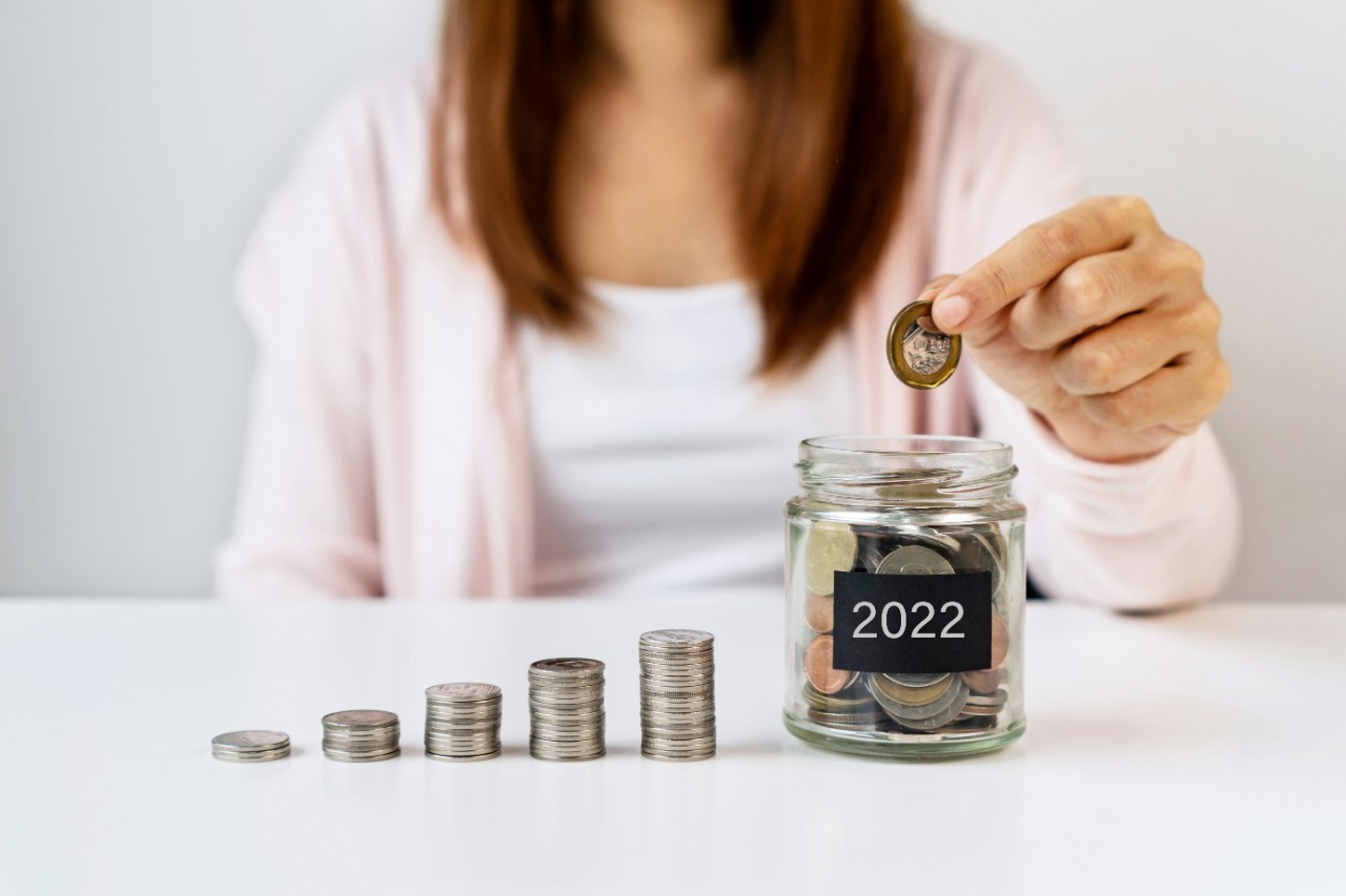 Hand of woman putting coin in glass jar labelled 2022 on white table background.