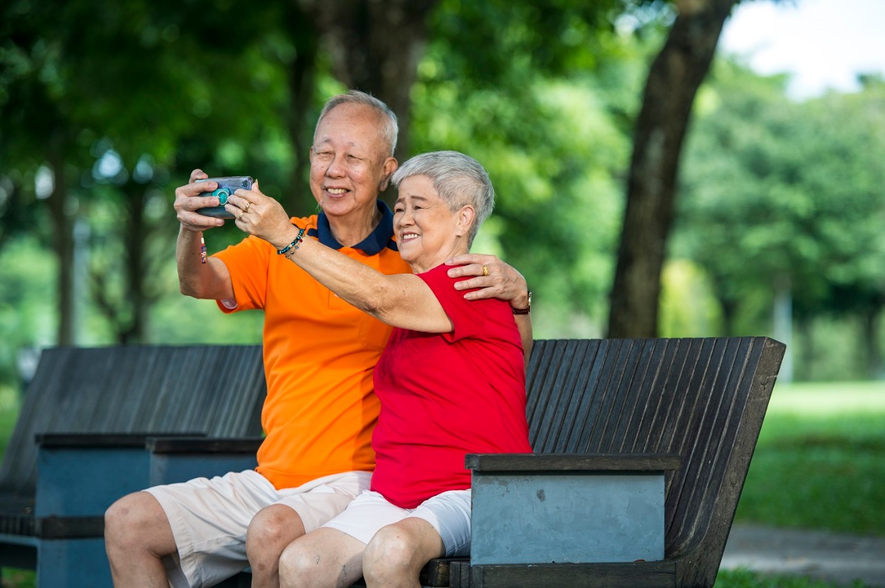 old elderly couple sitting on a park bench and taking a photo
