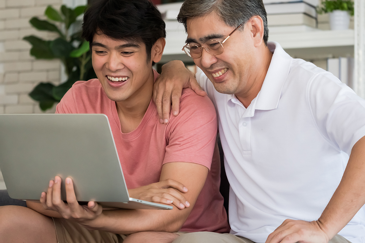 A senior male sits smiling beside his adult son, both looking at a laptop